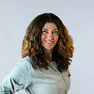 Eine Person mit lockigen, langen Haaren, die lächelt und eine helle Jacke trägt.